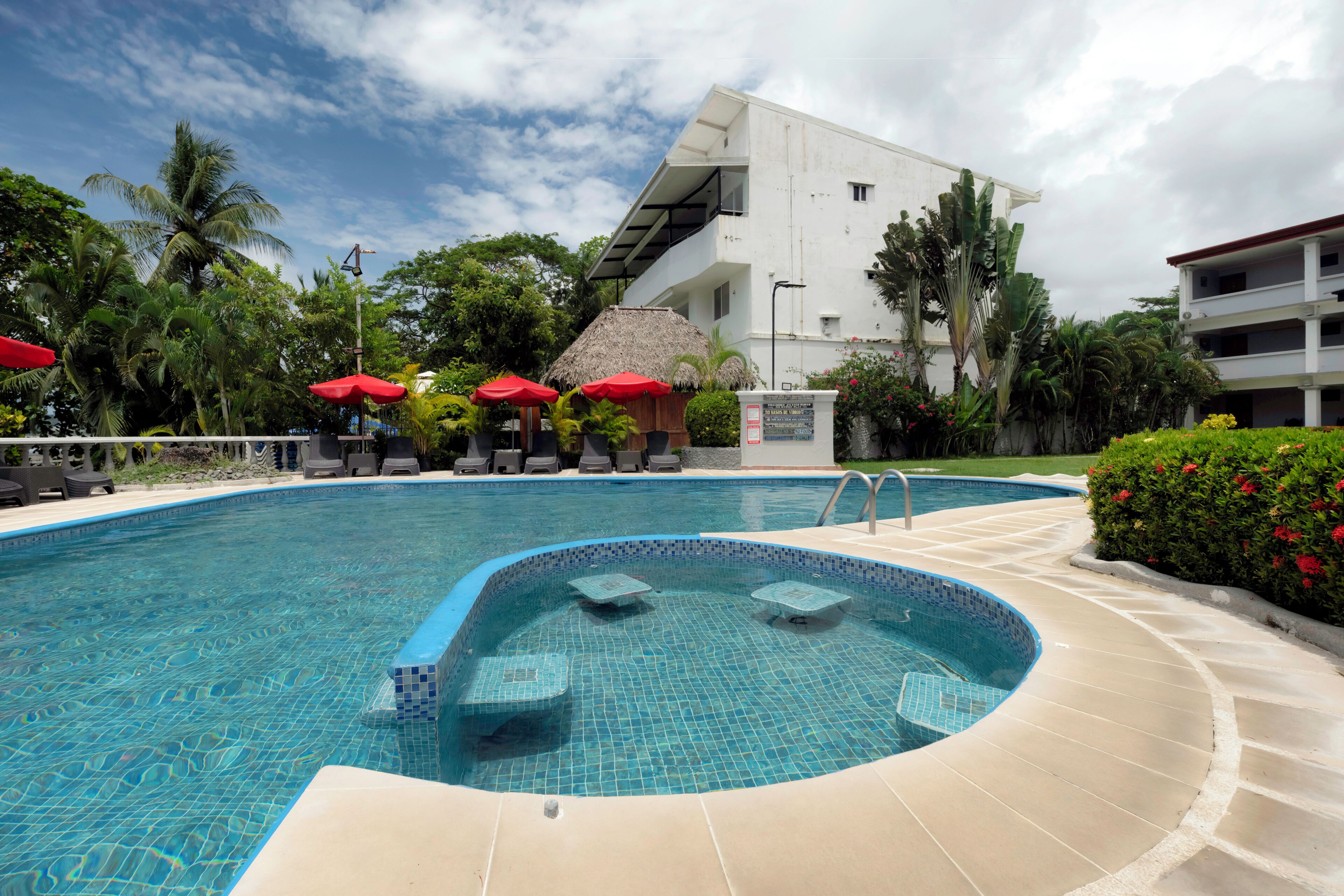 A swimming pool with a tiled edge, surrounded by lounge chairs and red umbrellas, with a building in the background.