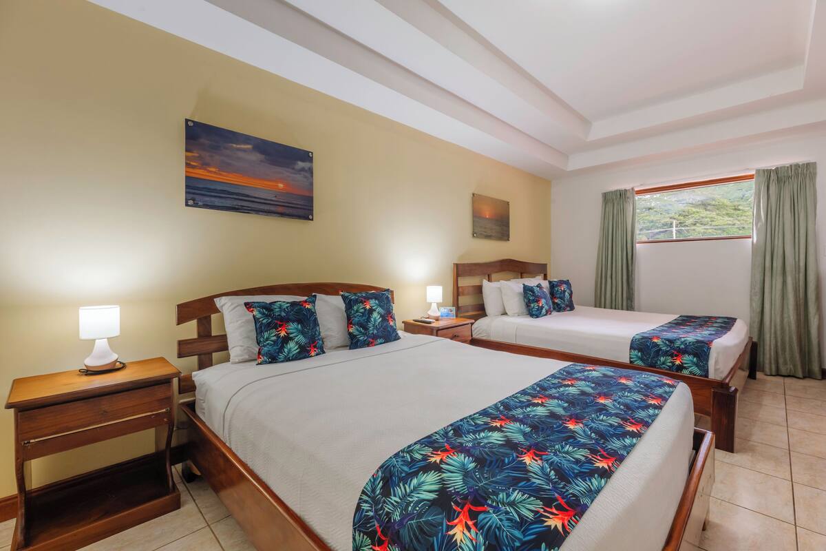 Habitación de hotel con dos camas, una mesita de noche con lámpara, un cuadro en la pared y una ventana con cortinas.