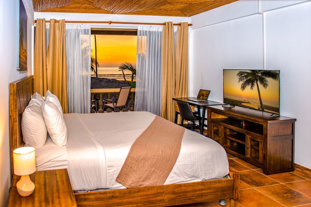 A hotel room with a bed, desk, TV, and a view of the sunset.