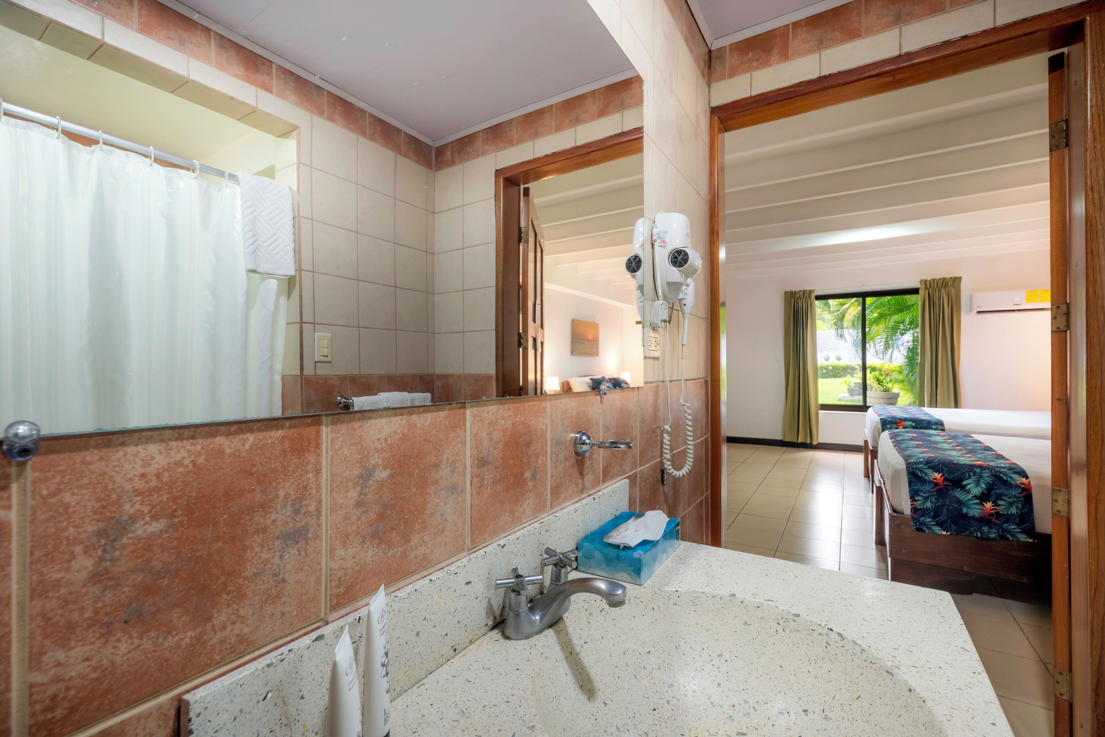 Un baño con un espejo grande, una ducha y un lavamanos. Hay un secador de pelo en la pared y una cama con una colcha estampada en la habitación contigua.