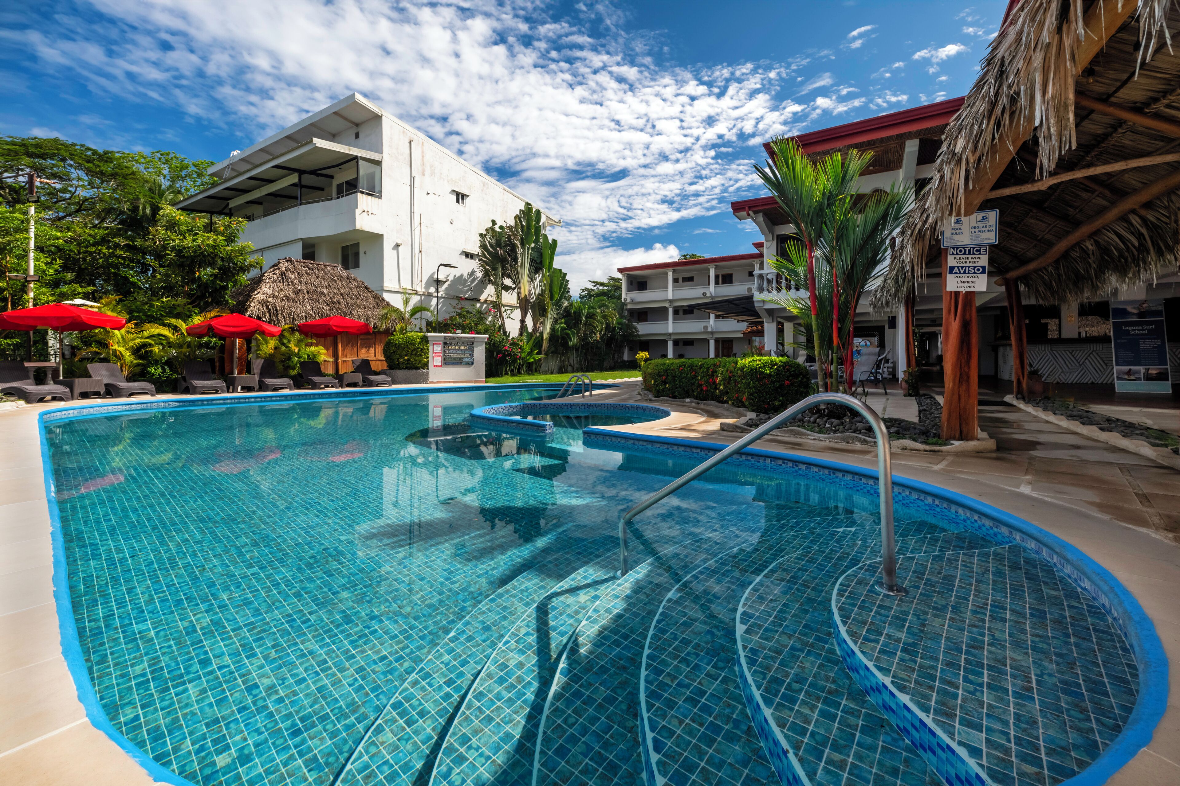 Ein Pool mit Fliesen umgeben, mit Liegestühlen und Sonnenschirmen, neben einem Gebäude mit einem Reetdach.