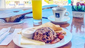 A breakfast plate with beans, sausage, toast, and a glass of orange juice on a wooden table with a view of the beach and ocean. - Jacó Laguna Resort and Beach Club (Jacó)