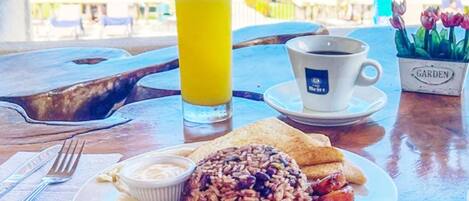 A breakfast plate with beans, sausage, toast, and a glass of orange juice on a wooden table with a view of the beach and ocean.