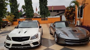 Two luxury cars parked in a driveway with a tiled floor and a fence in the background. - Tribeca Hotel (Accra)