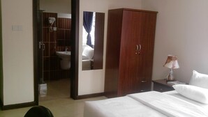 A hotel room with a bed, a wardrobe, a desk with a lamp, and a bathroom visible through an open door. - Tribeca Hotel (Accra)