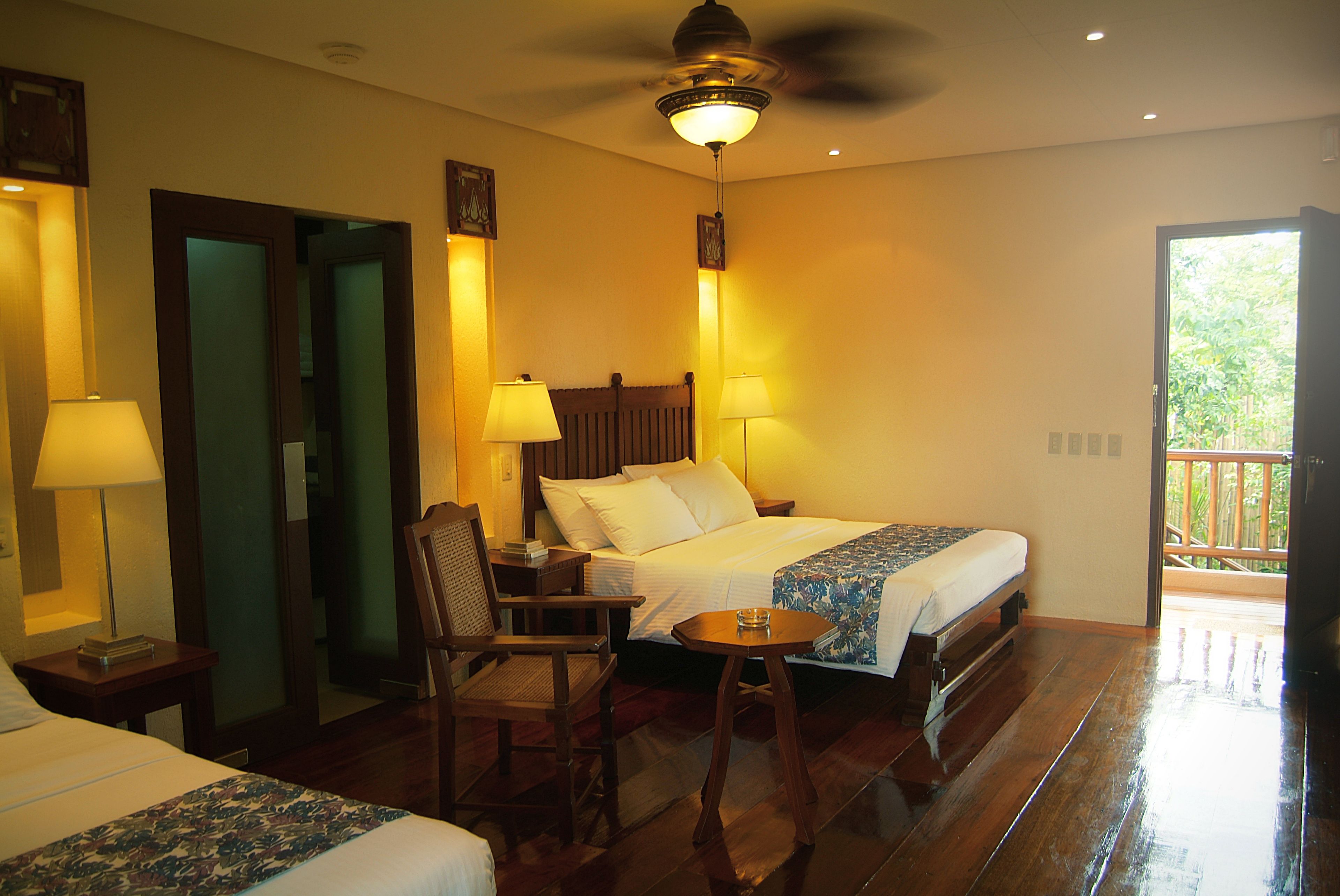 A hotel room with a bed, two bedside lamps, a chair, a small table, and a ceiling fan.
