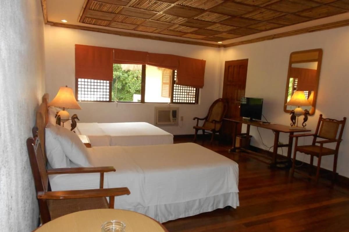 A room with two beds, wooden furniture, a desk with a computer, and a window with a view of greenery.
