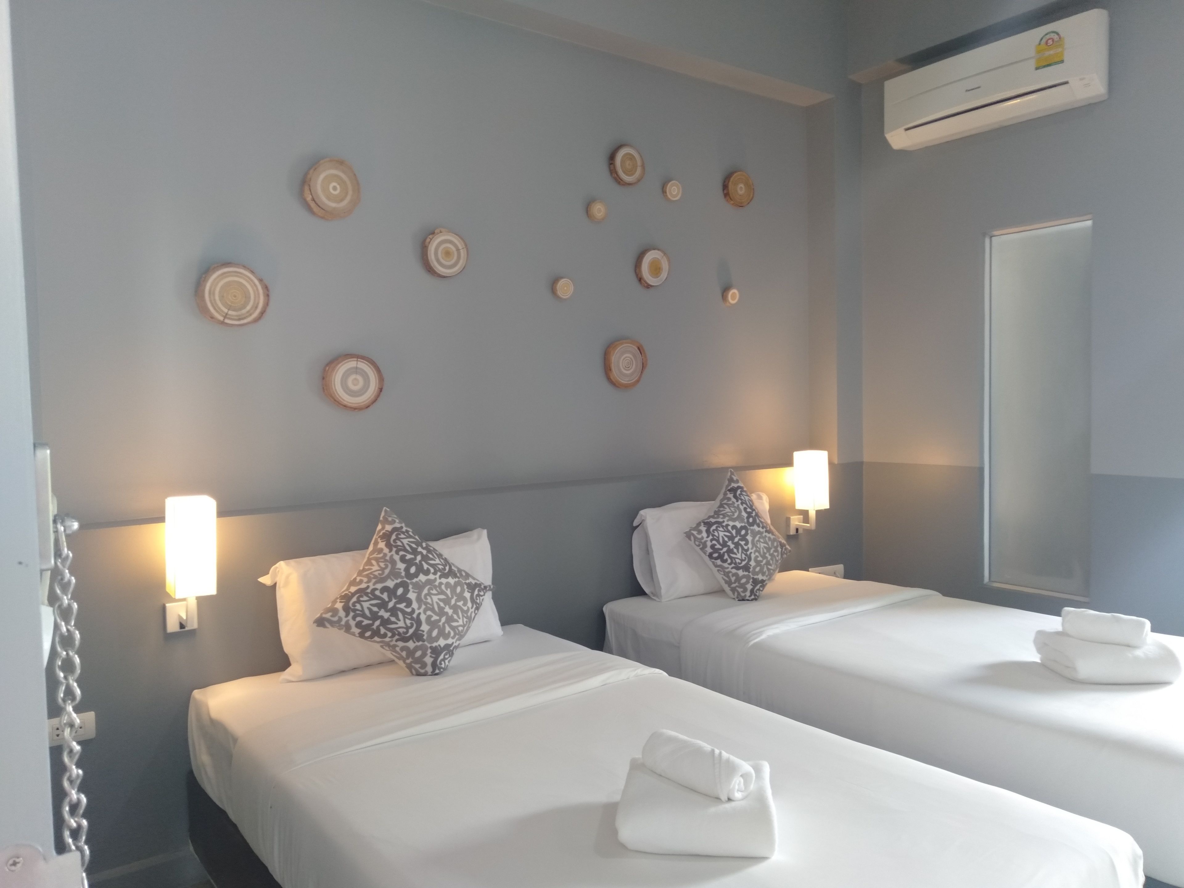 Habitación de hotel con dos camas, ropa blanca, almohadones estampados, lámparas de pared y platos de madera decorativos.