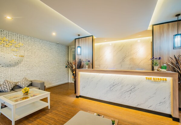 A reception area with a marble counter, a sofa with patterned cushions, and a coffee table with flowers. - The Rubber Hotel Phuket (Thep Krasattri)