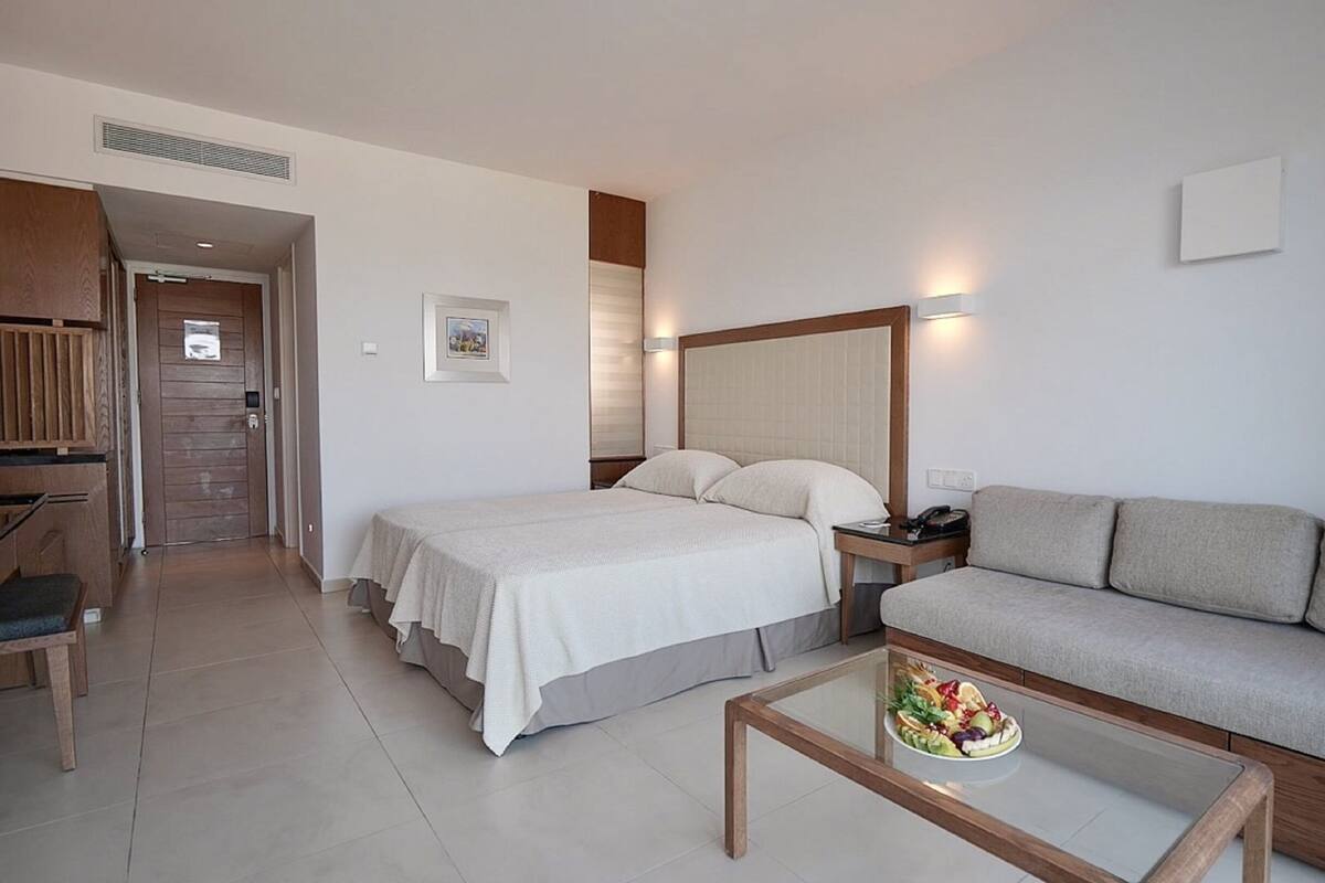 Une chambre d’hôtel comprenant un lit, un canapé, une table basse en verre avec des fruits, un bureau et une porte donnant sur une autre pièce.