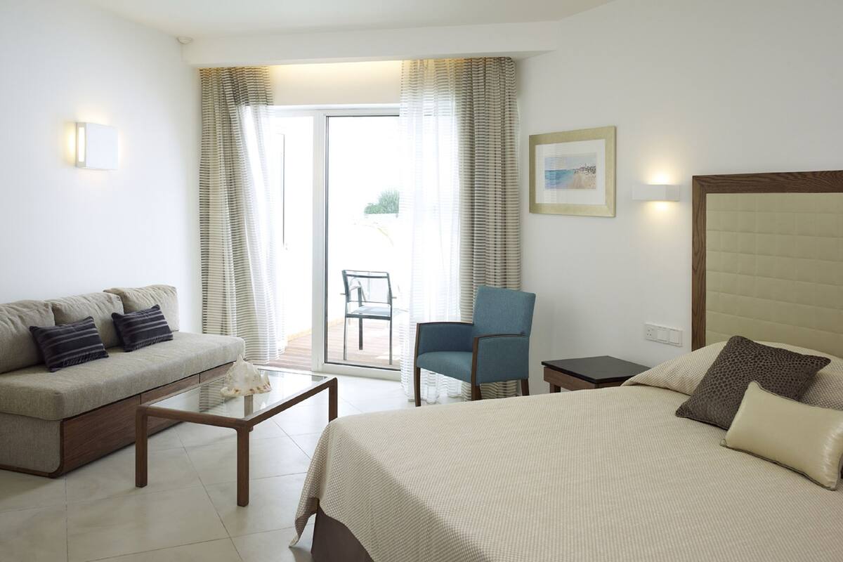 Une chambre d’hôtel avec un lit, un canapé, une chaise, une petite table et une vue sur l’extérieur, visible à travers une porte vitrée.