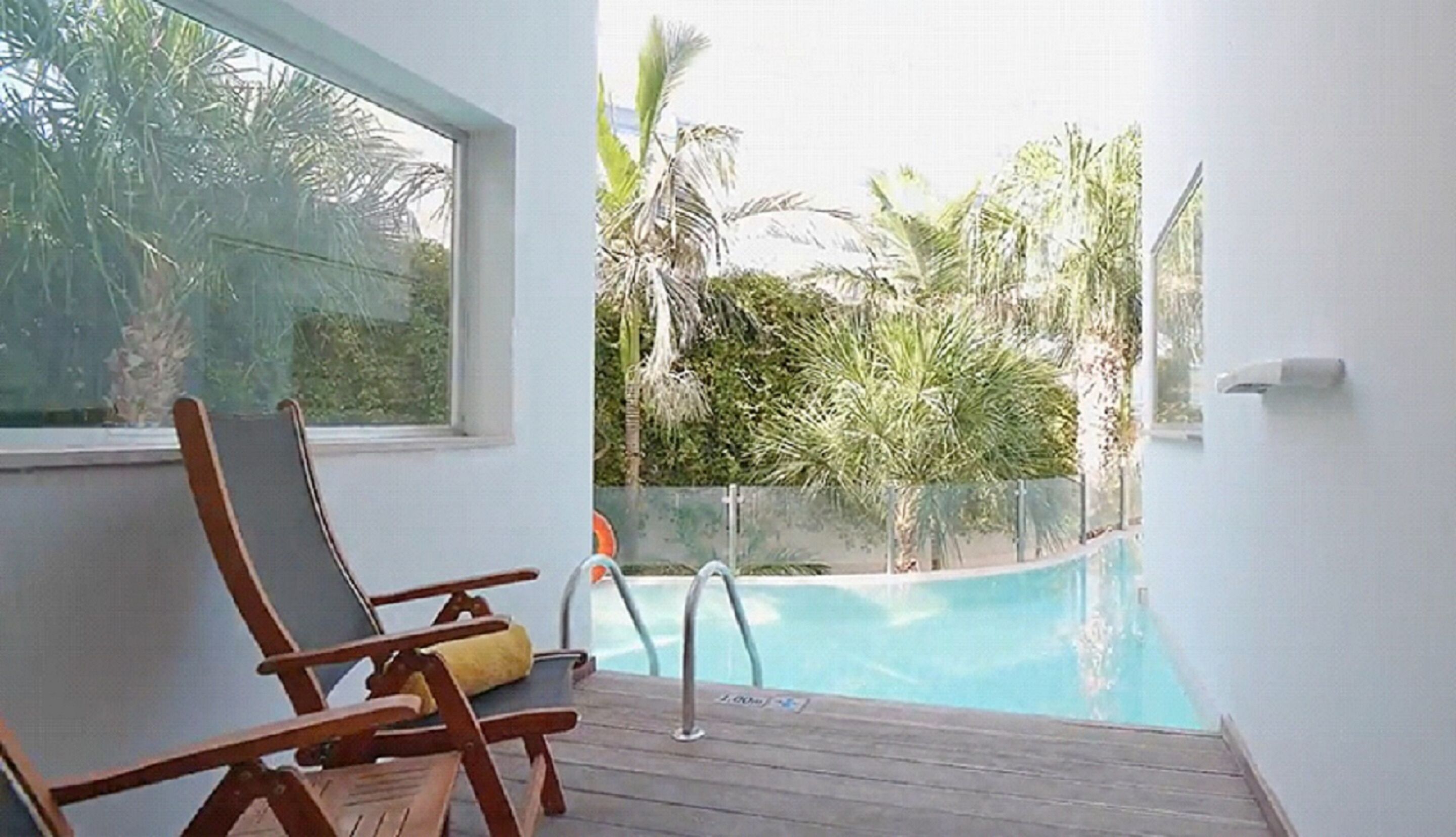 A poolside area with a wooden deck, a lounge chair, and a view of a swimming pool surrounded by tropical vegetation.