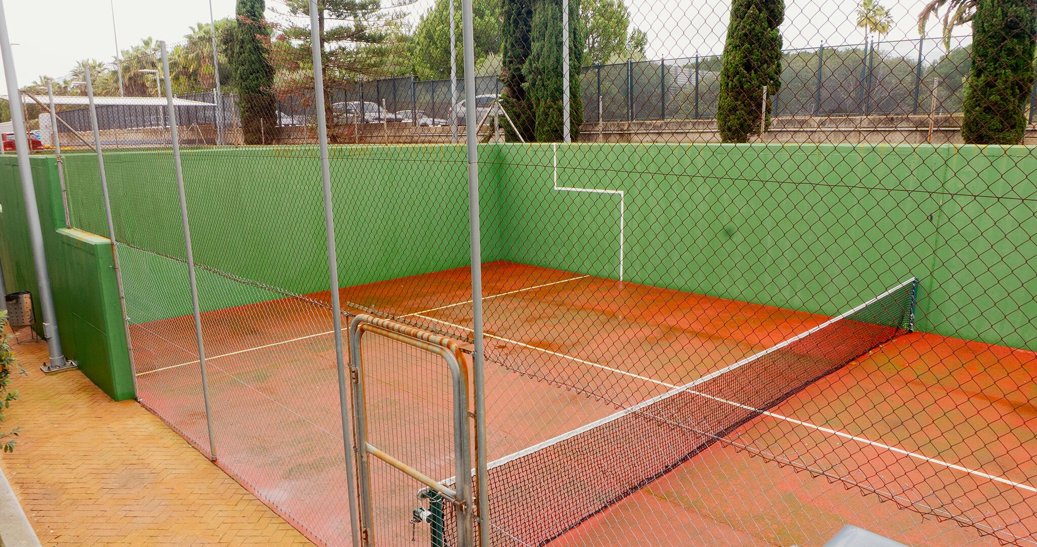 Una cancha de tenis con superficie verde y roja, rodeada por una cerca de malla metálica.