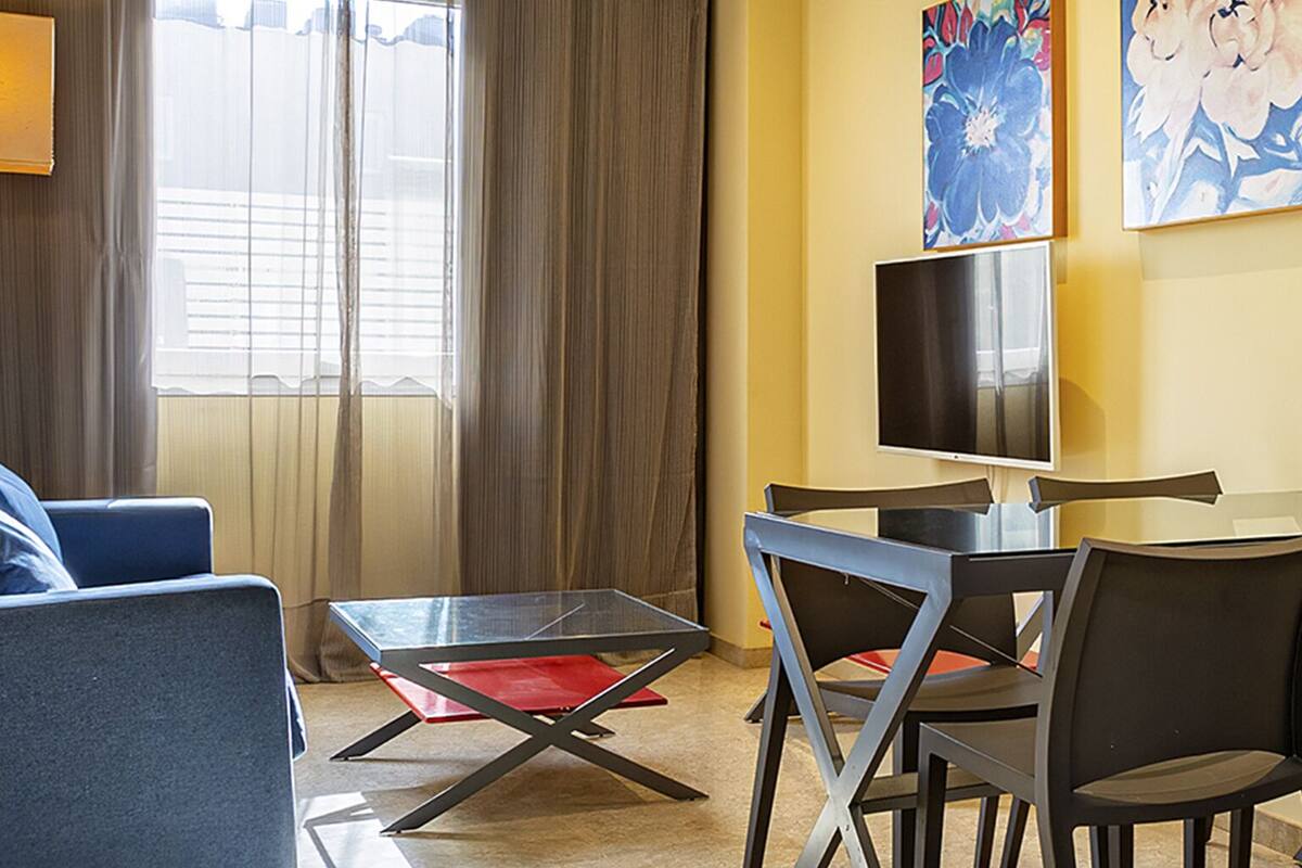Ein modernes Hotelzimmer mit einer Couch, einem kleinen Tisch, einem Fernseher und einem Fenster mit Vorhängen.
