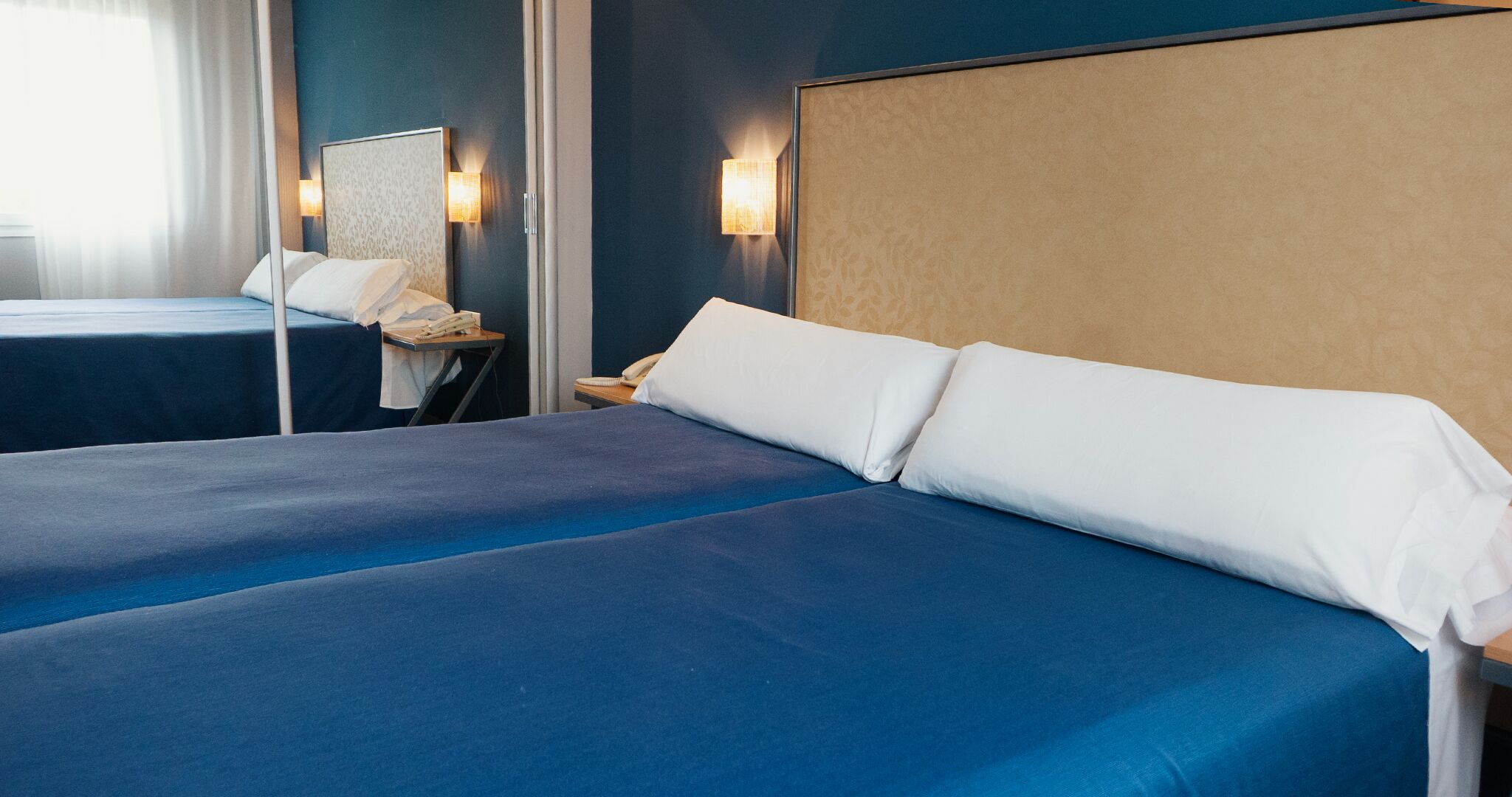 Une chambre d’hôtel avec deux lits, chacun recouvert d’une couverture bleue et doté d’oreillers blancs. Il y a une table de chevet avec une lampe et un téléphone.