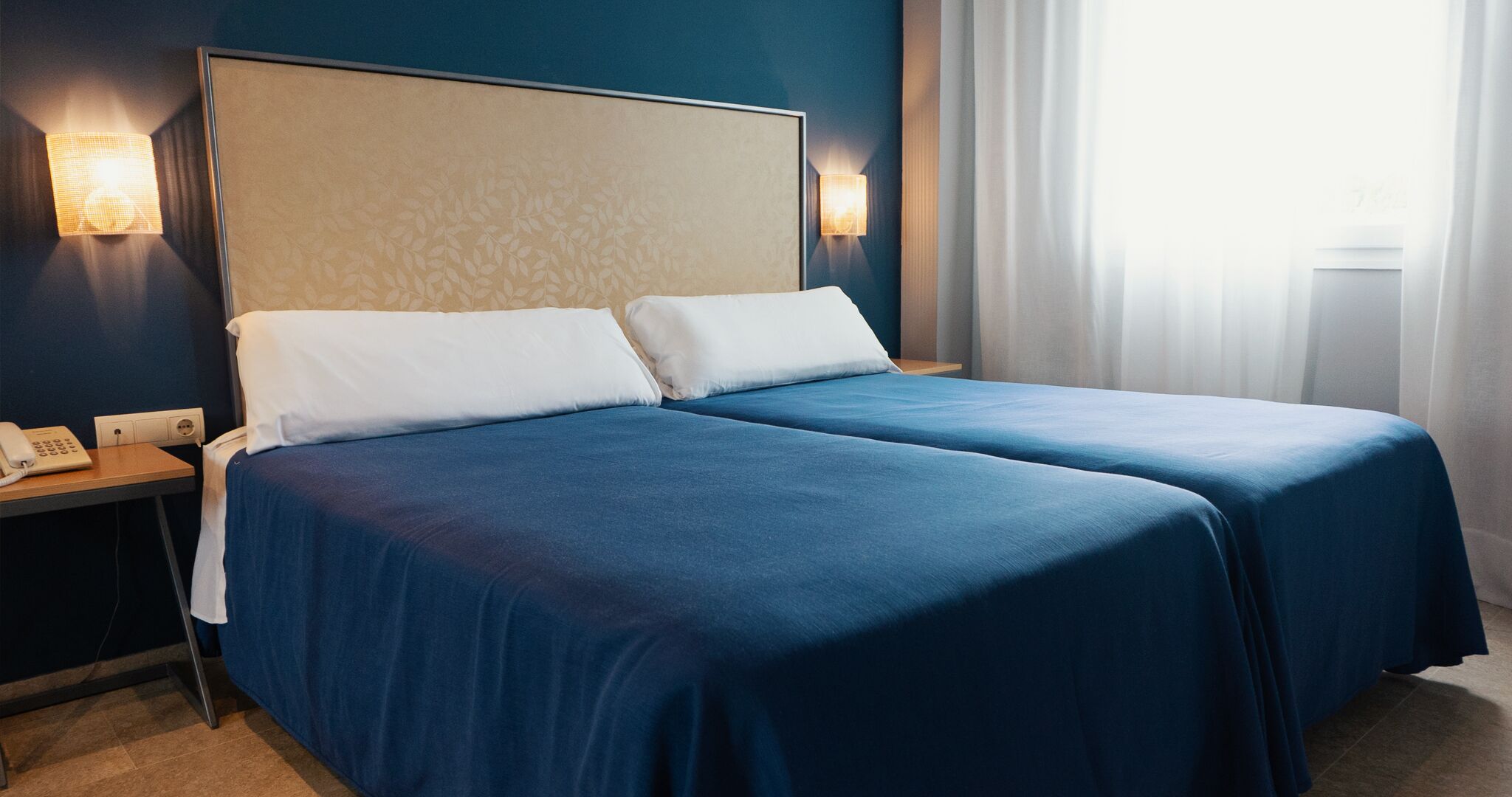 A hotel room with two beds, a nightstand with a phone, and wall-mounted lights.