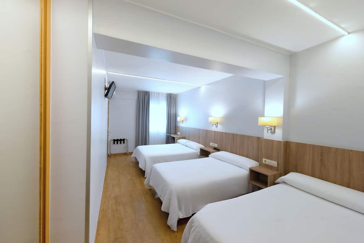 A hotel room with three single beds, a wooden headboard, bedside tables with lamps, a flat-screen TV, and a window with curtains.