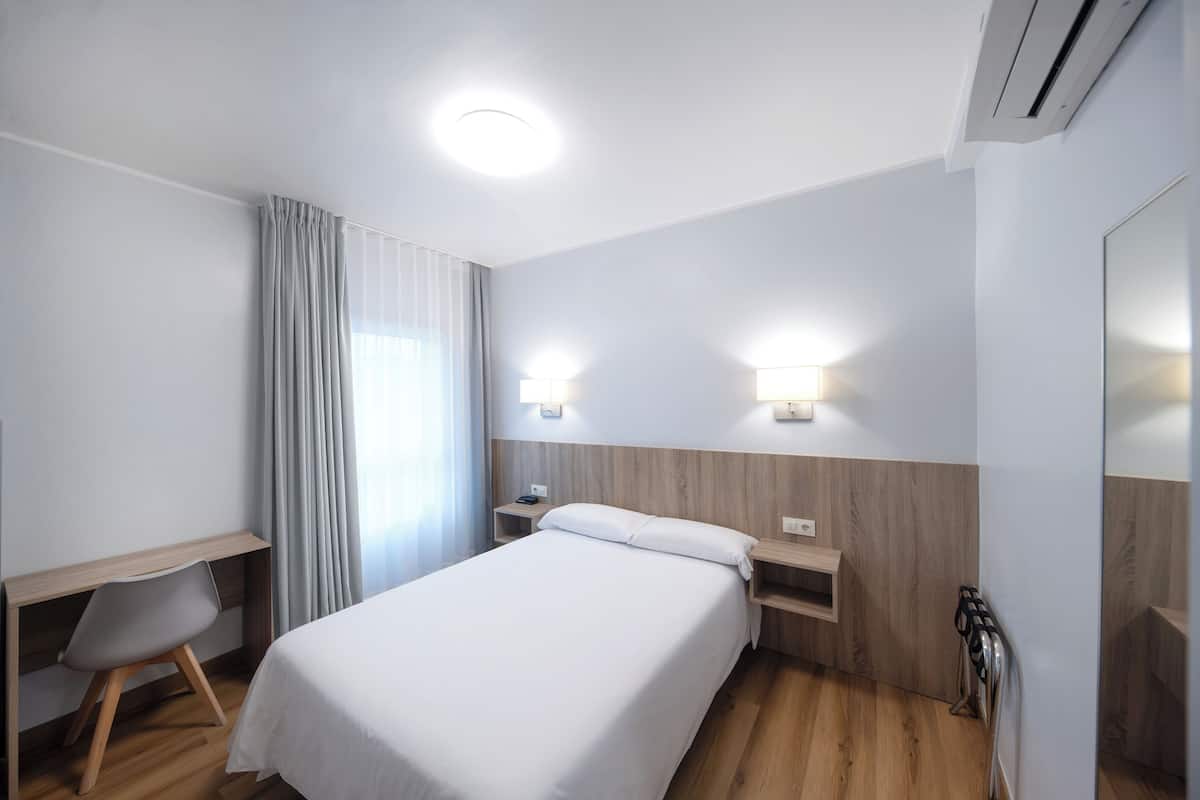 A hotel room with a bed, a desk, and a chair. There is a nightstand with a lamp, a window with curtains, and a wall-mounted radiator.