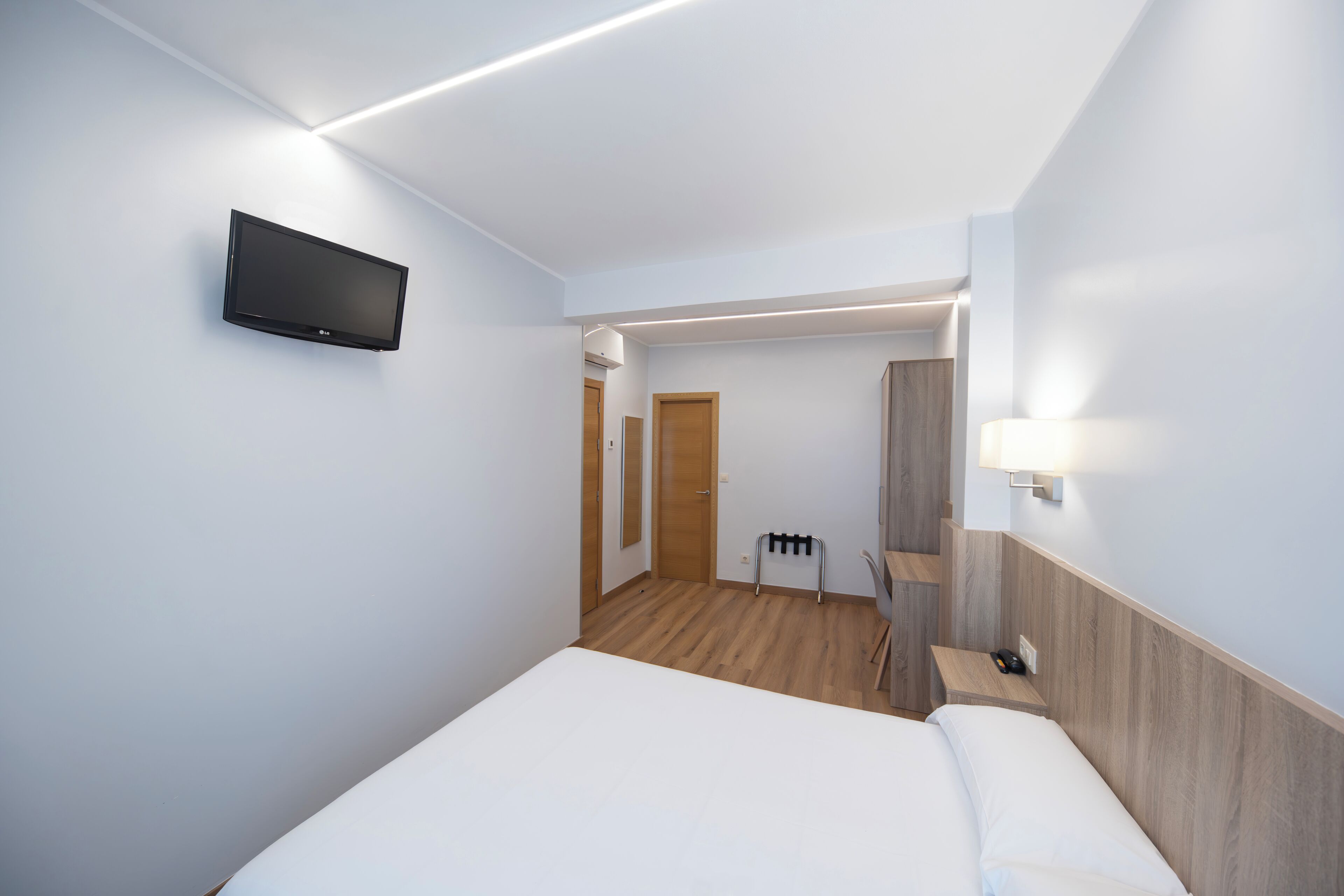 Habitación de hotel con cama, televisor montado en la pared, suelo de madera, una puerta y una mesita con lámpara.