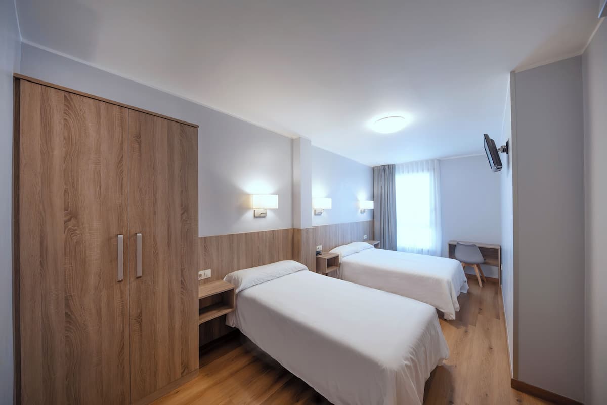 A hotel room with two single beds, a wooden wardrobe, bedside tables, and a desk with a chair.