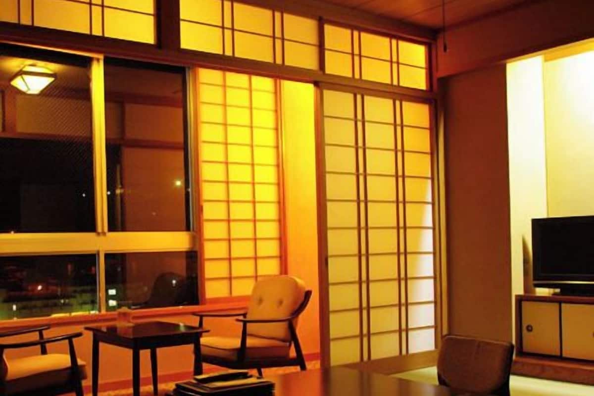 A room with tatami flooring, a low table, a sofa, a TV on a cabinet, and large windows with a view of the outside.