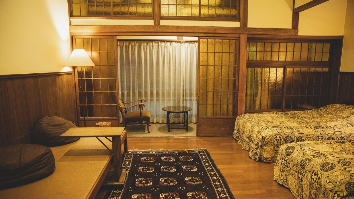 Una habitación tradicional japonesa con suelo de tatami, una mesa baja con cojines, una cama futón, una mesita redonda y un puertas correderas con cortinas.