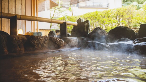 Un área de aguas termales con rocas, vapor, estructuras de madera y follaje al fondo.