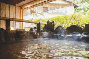 Un área de aguas termales con rocas, vapor, estructuras de madera y follaje al fondo.