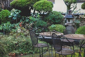 A garden patio with metal chairs and a round table, surrounded by lush greenery and a stone lantern.