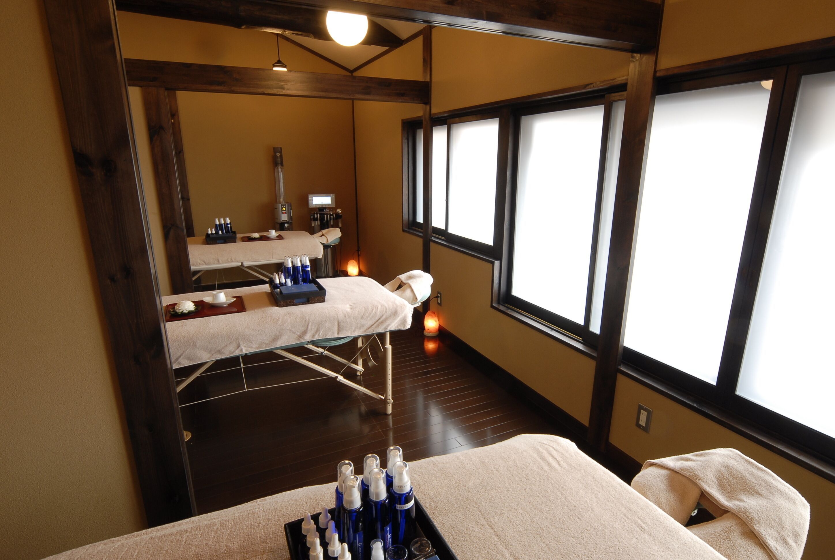 A room with a massage table, a window with blinds, a tray with bottles, and a small table with a bowl and cups.