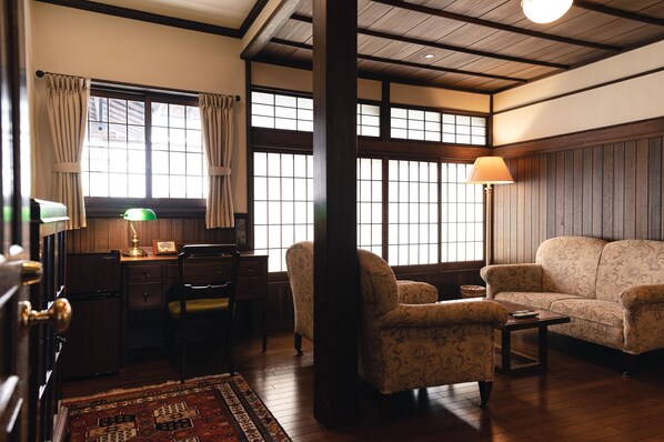 A traditional room with wooden floors, a sofa, a chair, a desk with a lamp, and large windows with curtains.