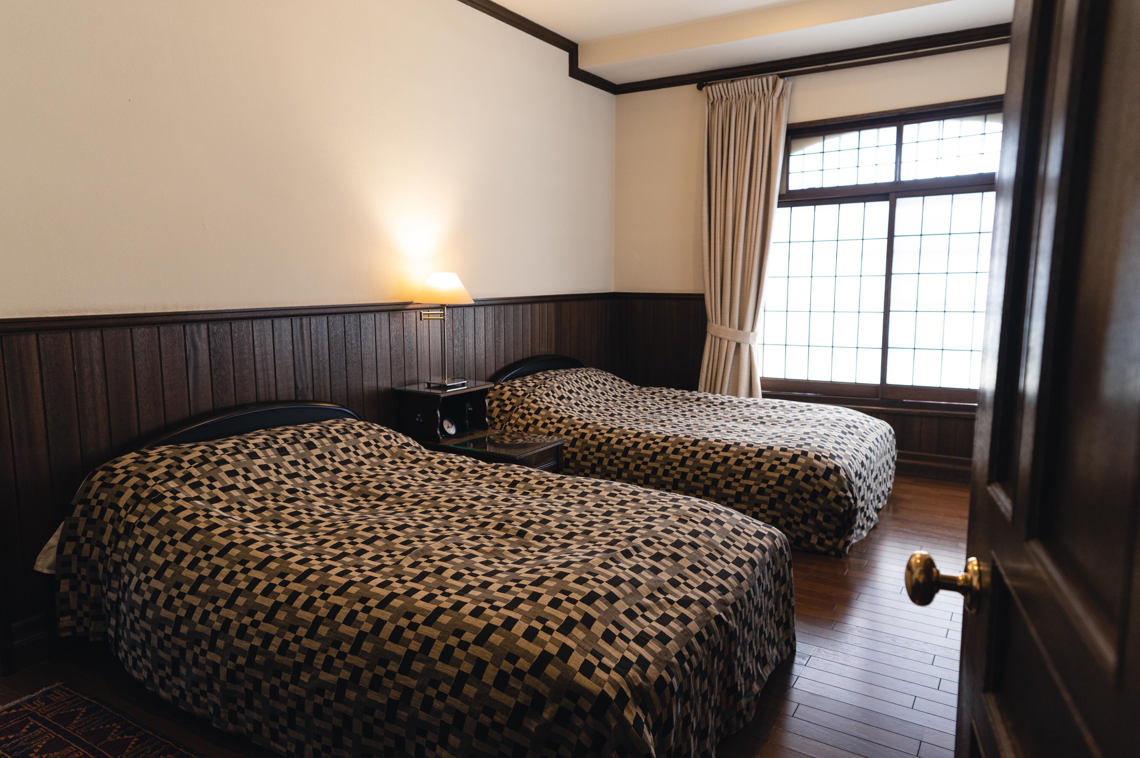 A room with two beds, a nightstand, a lamp, and a window with curtains.