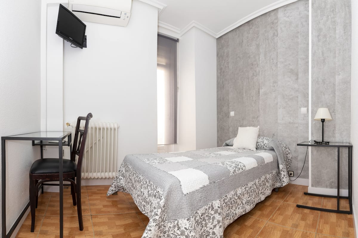 A bedroom with a bed, a desk, a chair, a lamp, and a TV mounted on the wall.