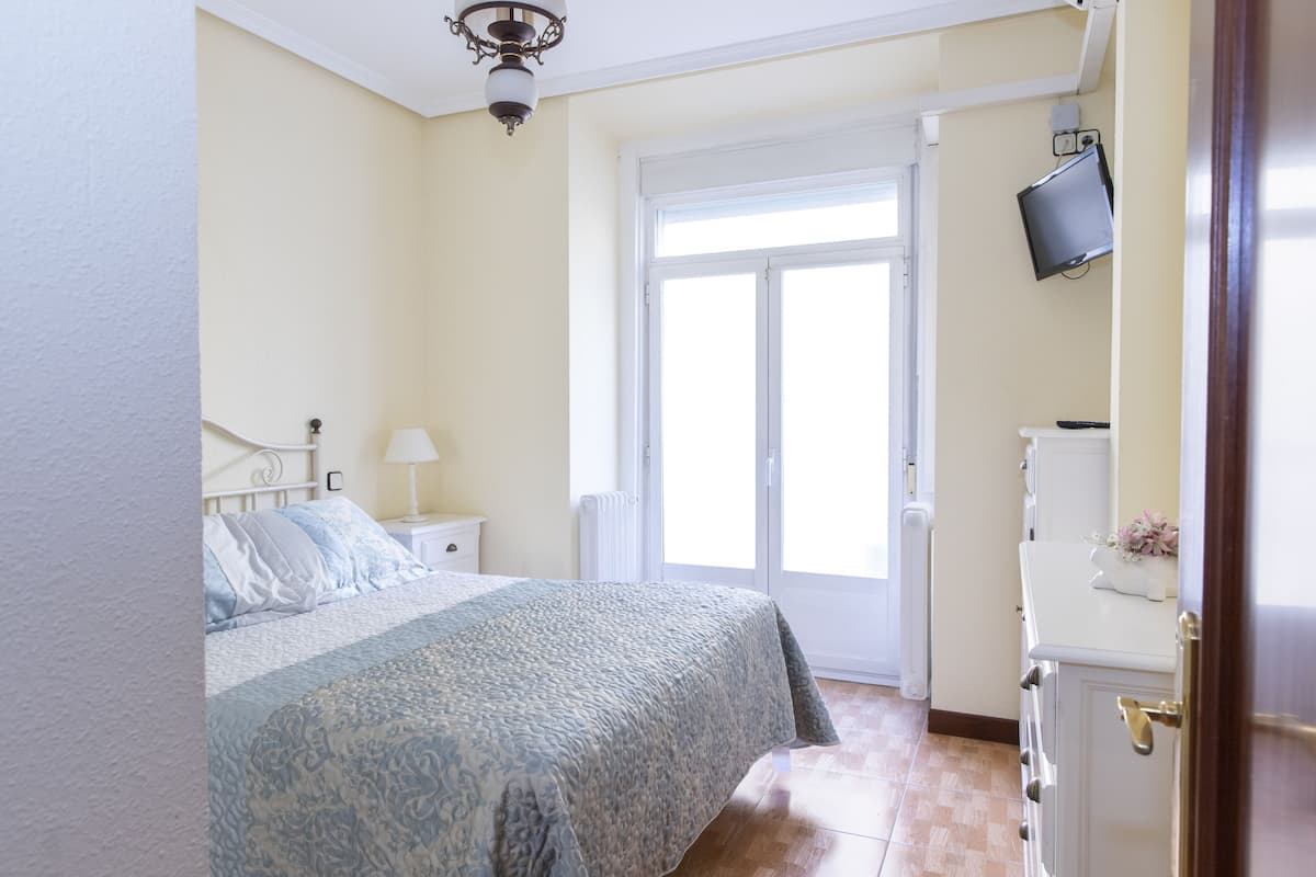 A bedroom with a bed, a nightstand, a lamp, a television on the wall, and a door leading to a balcony.