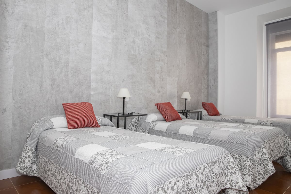 A hotel room with two beds, grey floral bedding, red pillows, bedside tables with lamps, and a concrete wall.