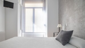 Ein Hotelzimmer mit einem großen Bett, einem flachbildfernseher an der Wand, einem Fenster mit Jalousien und einem Nachttisch mit Lampe.
