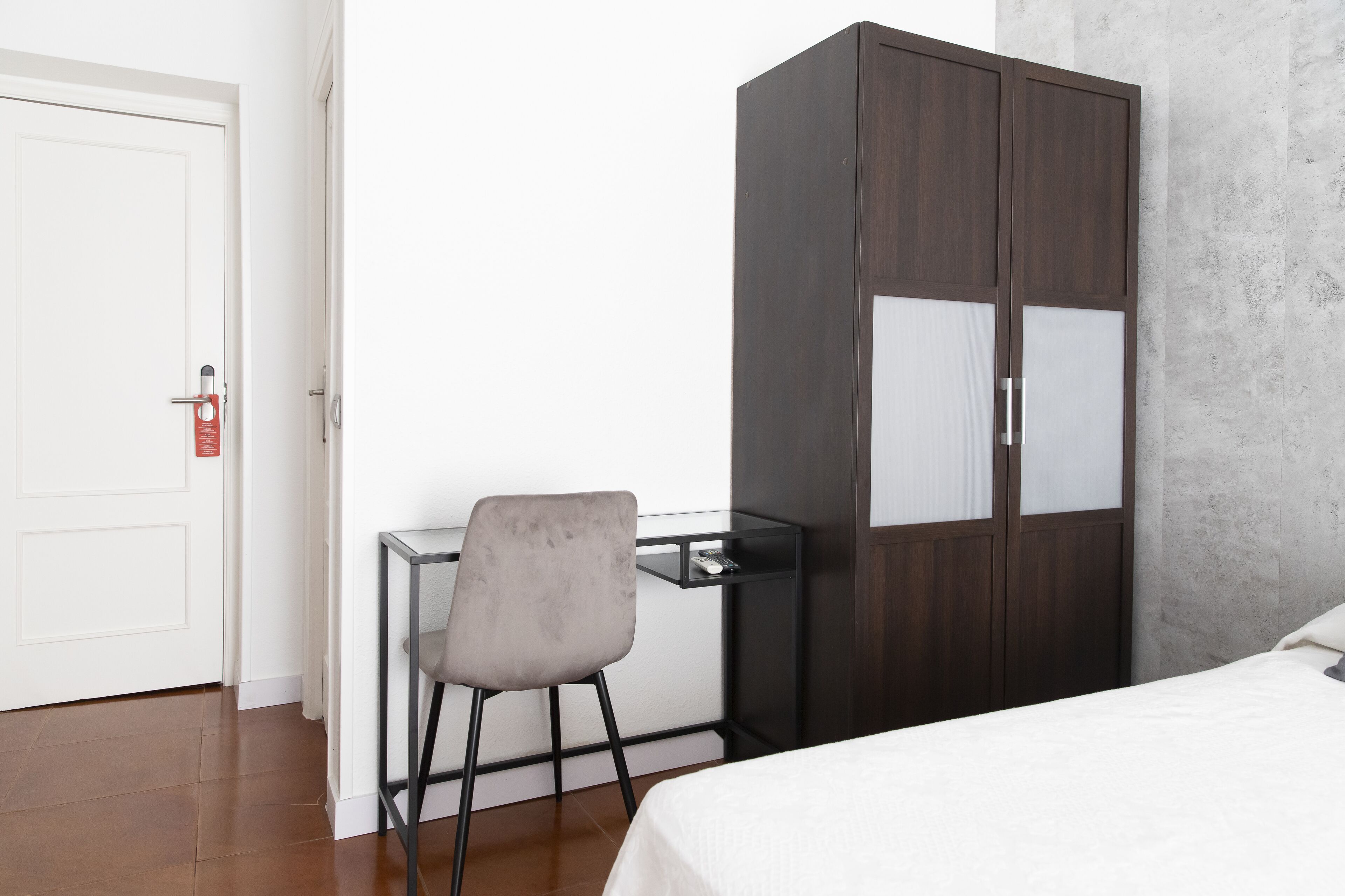 Un dormitorio con una cama, un escritorio con silla, un armario y una puerta con detector de incendios.