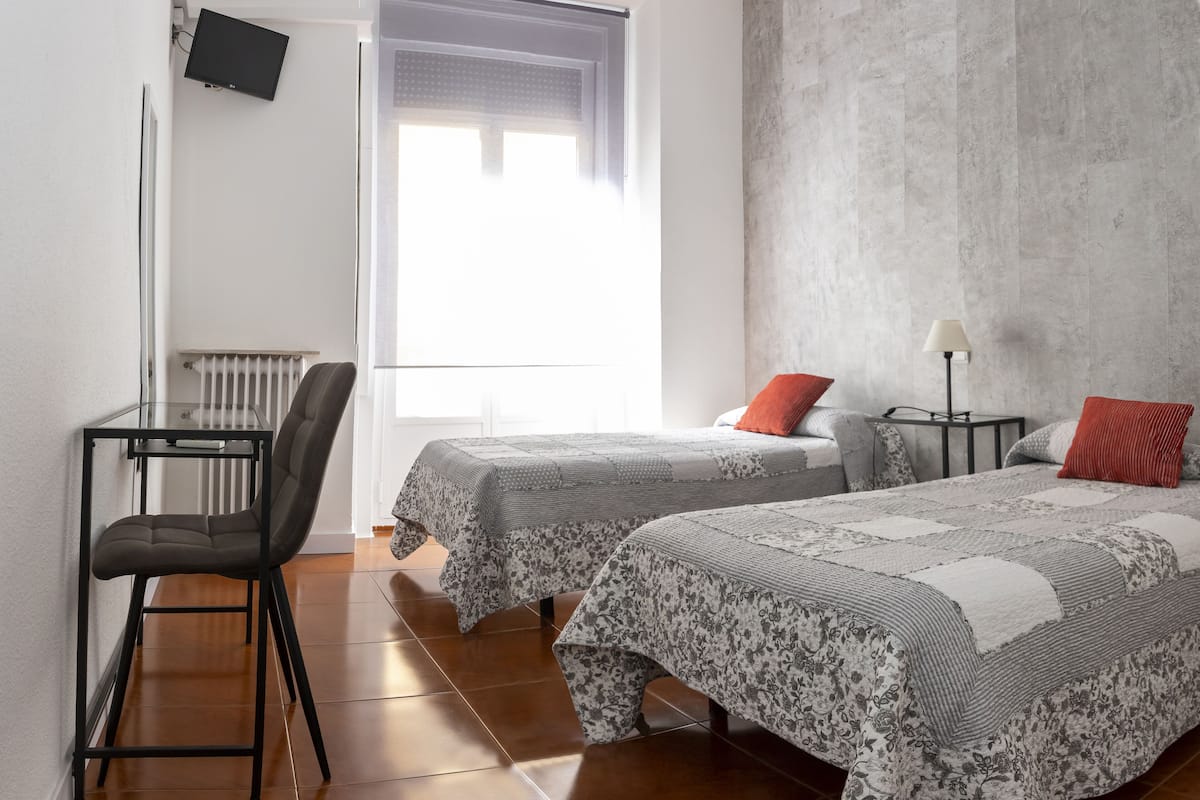 A room with two beds, a desk, a chair, a TV, and a window with blinds.