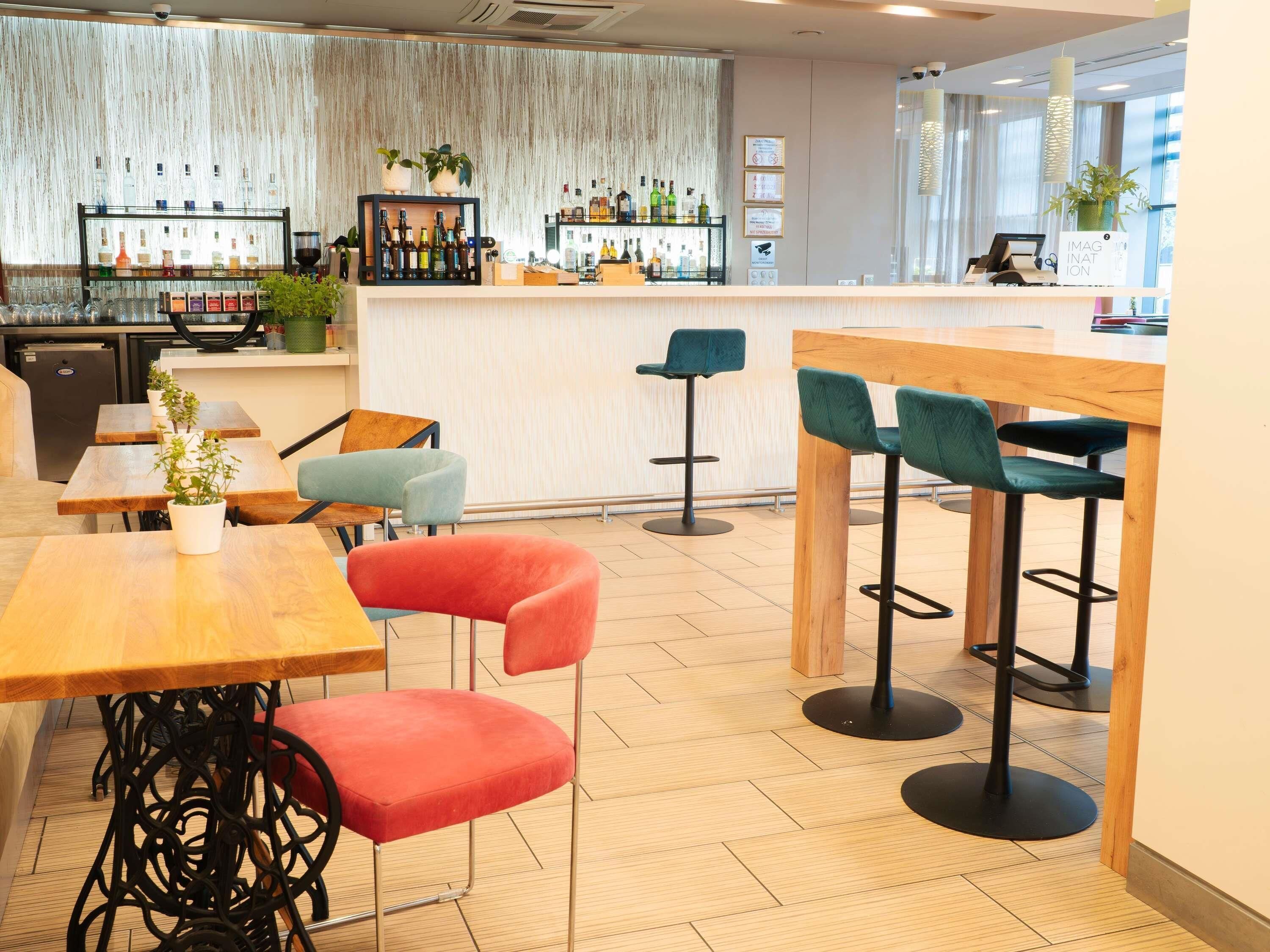 A modern bar area with wooden tables and colorful bar stools, a bar counter with various bottles, and a large window providing natural light.
