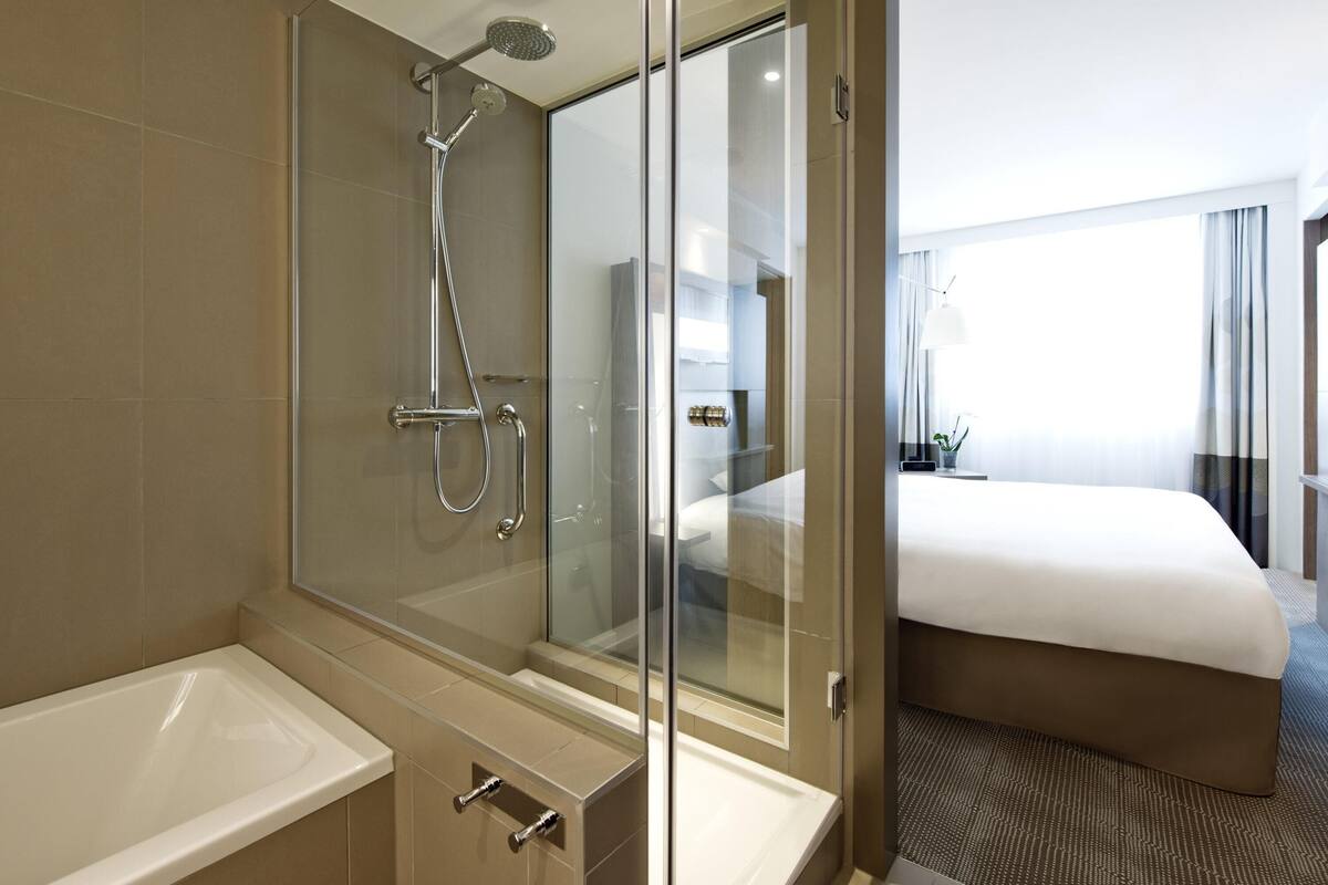 A modern hotel bathroom with a glass-enclosed shower, a bathtub, and a visible bedroom with a bed and a nightstand.