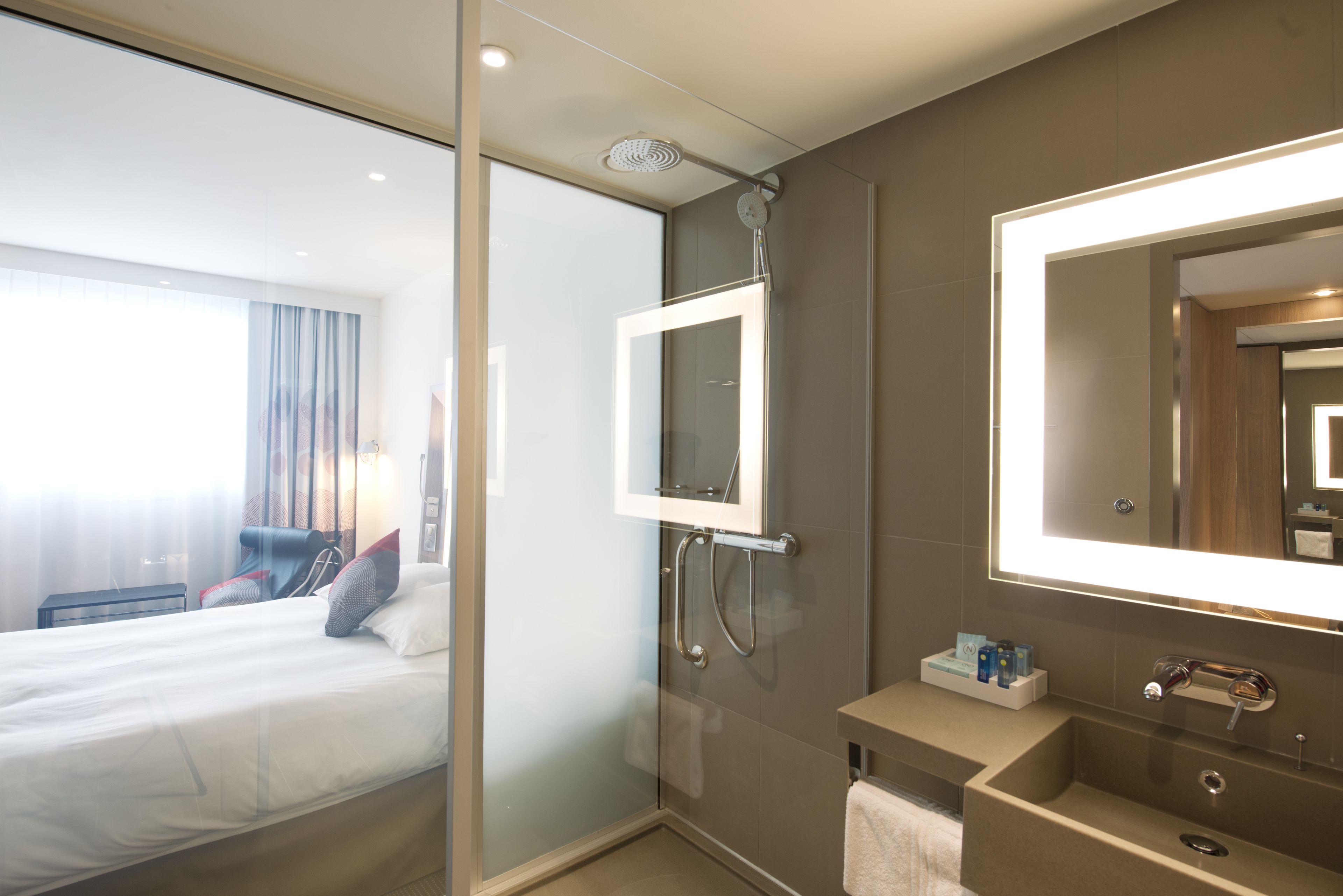 Habitación de hotel con cama, cabina de ducha de vidrio, tocador con espejo y una pequeña estantería con artículos de aseo.