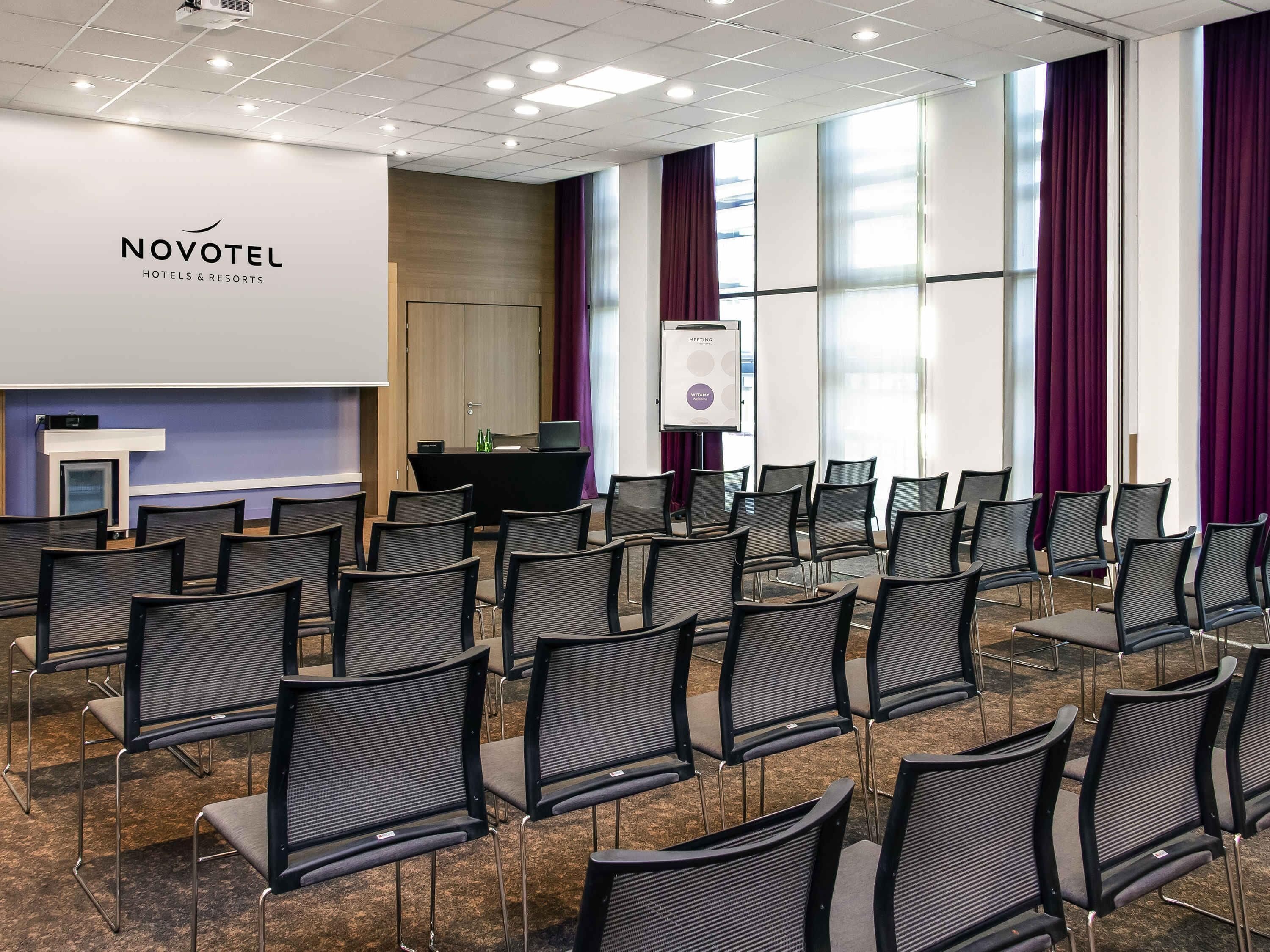 Un salón de conferencias con filas de sillas, un atril, una pantalla para presentaciones y un cartel que anuncia una reunión.