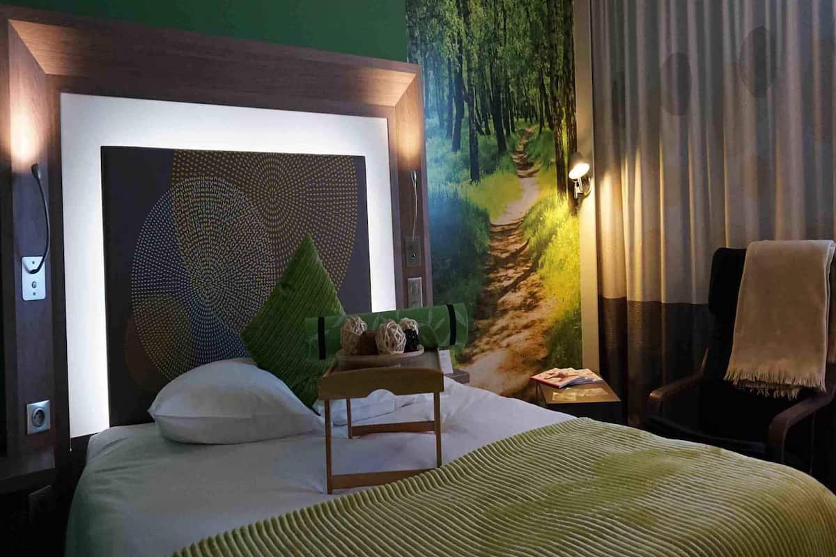A hotel room with a large bed, a headboard with a geometric pattern, a green wall mural, a small table with a lamp, and a chair with a blanket.