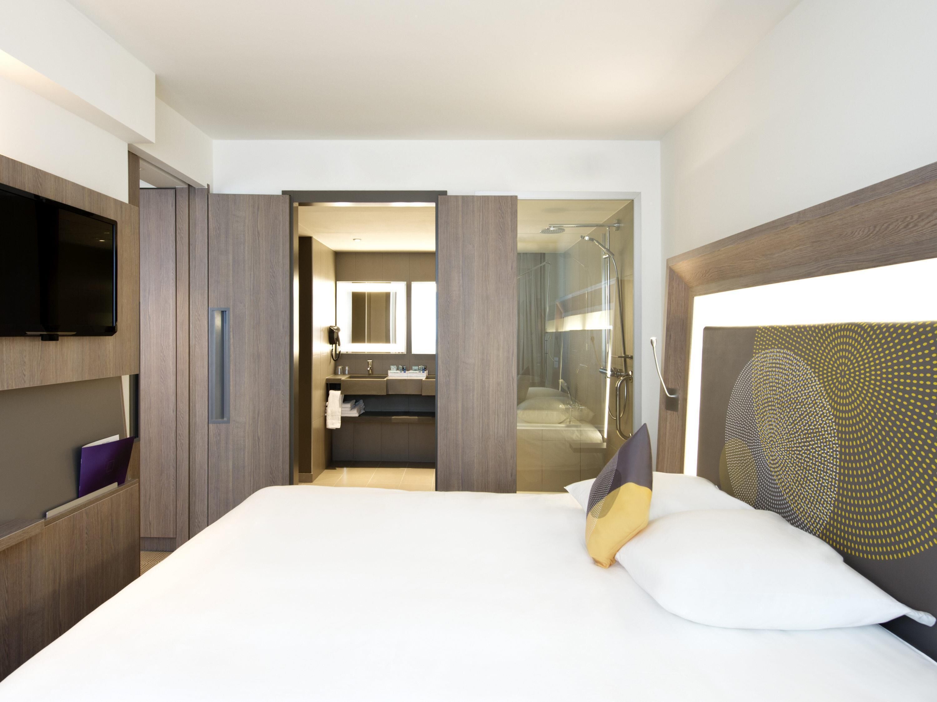 A modern hotel room with a large bed, a TV, a bathroom with a shower, and a walk-in closet.