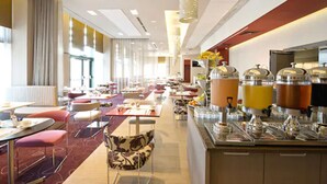 A modern hotel dining area with multiple tables and chairs, a buffet station with various food containers, and a large window providing natural light. - Novotel Lodz Centrum (Lodz)