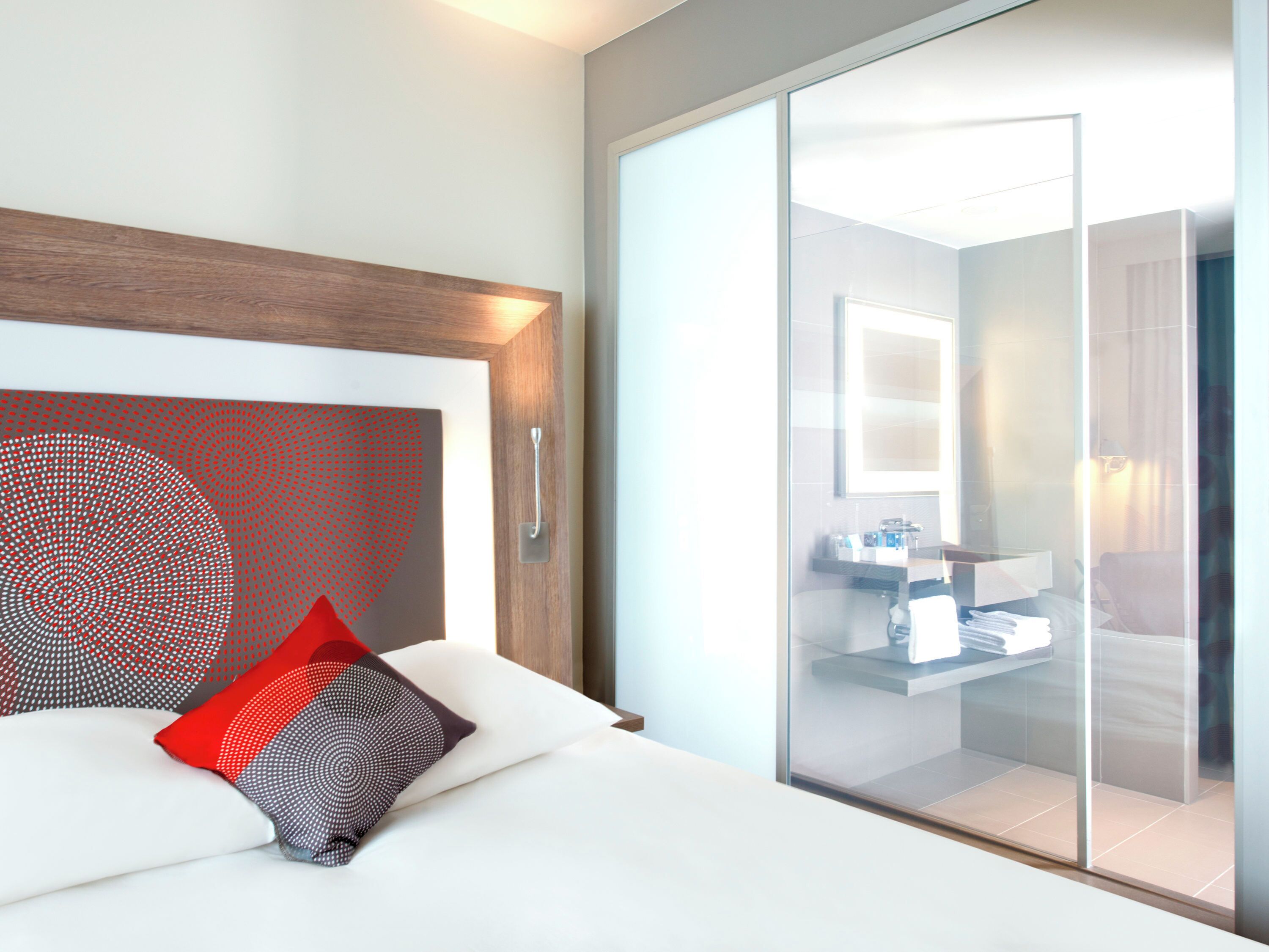A modern hotel room with a bed, a large mirror, and a sliding glass door leading to a bathroom.
