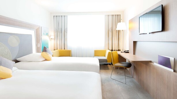 A hotel room with two beds, a desk, a chair, a TV, and a window with curtains. - Novotel Lodz Centrum (Lodz)