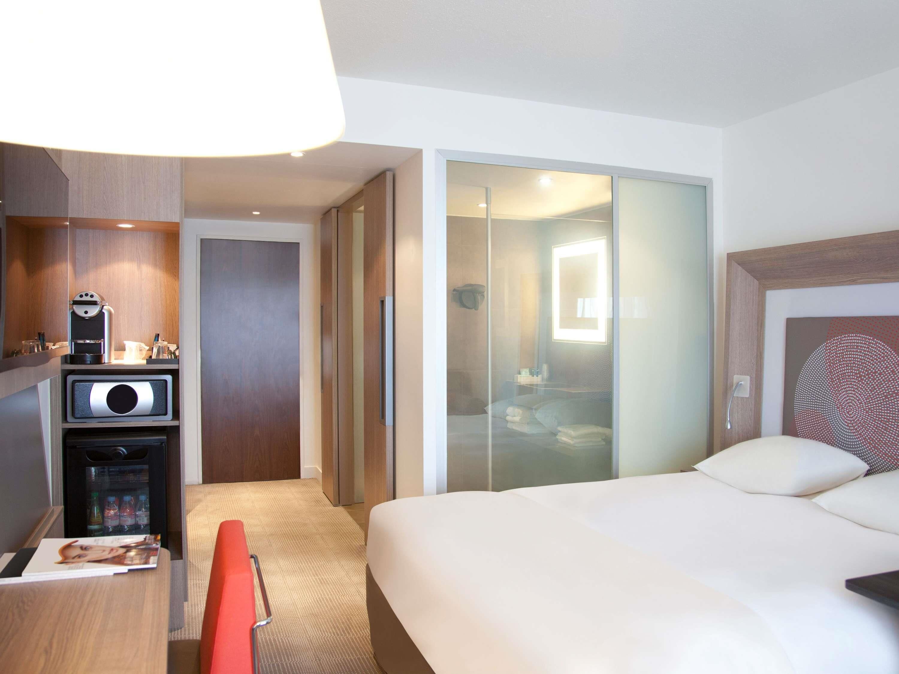 A hotel room with a bed, a desk with a coffee machine, a mini-fridge, and a sliding door leading to a bathroom.