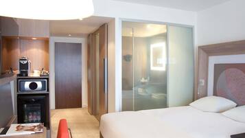 A hotel room with a bed, a desk with a coffee machine, a mini-fridge, and a sliding door leading to a bathroom.