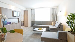A modern hotel room with a grey sofa, a small dining table, a coffee table, and a potted plant. - Novotel Lodz Centrum (Lodz)